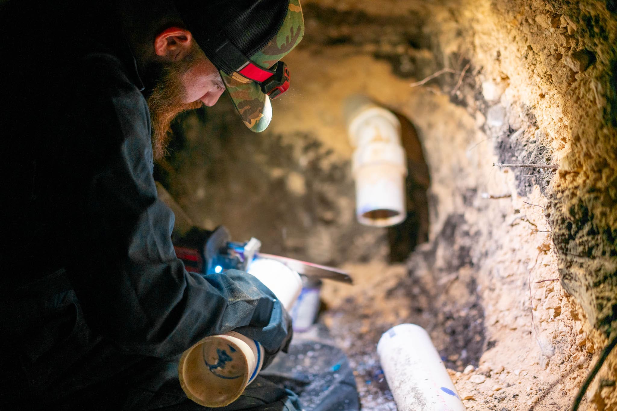 The Bearded Plumber technician working on pipe beneath a foundation